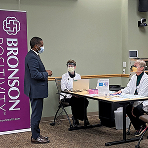 Bill manns standing in front of masked individuals at a table