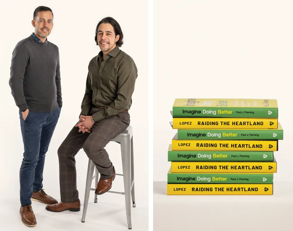 Paul Fleming standing to left with Bill Lopez sitting atop a stool. To the right, a stack of their books, eight total, alternating with Fleming's "Imagine Doing Better" in green and then Lopez's "Raiding the Heartland" in Yellow, and so on.  