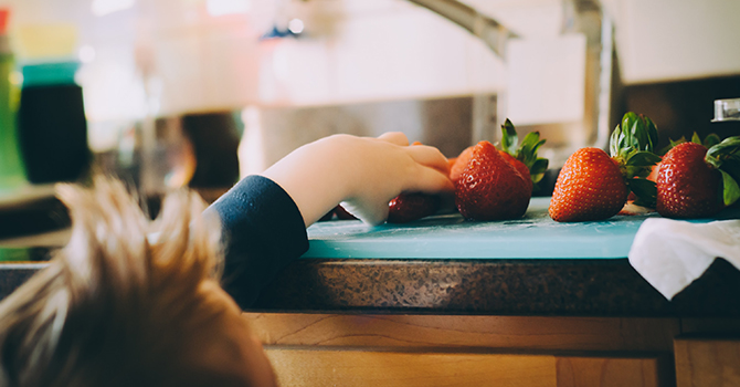 Young child reaching for strawberries on a cutting board on a kitchen counter.