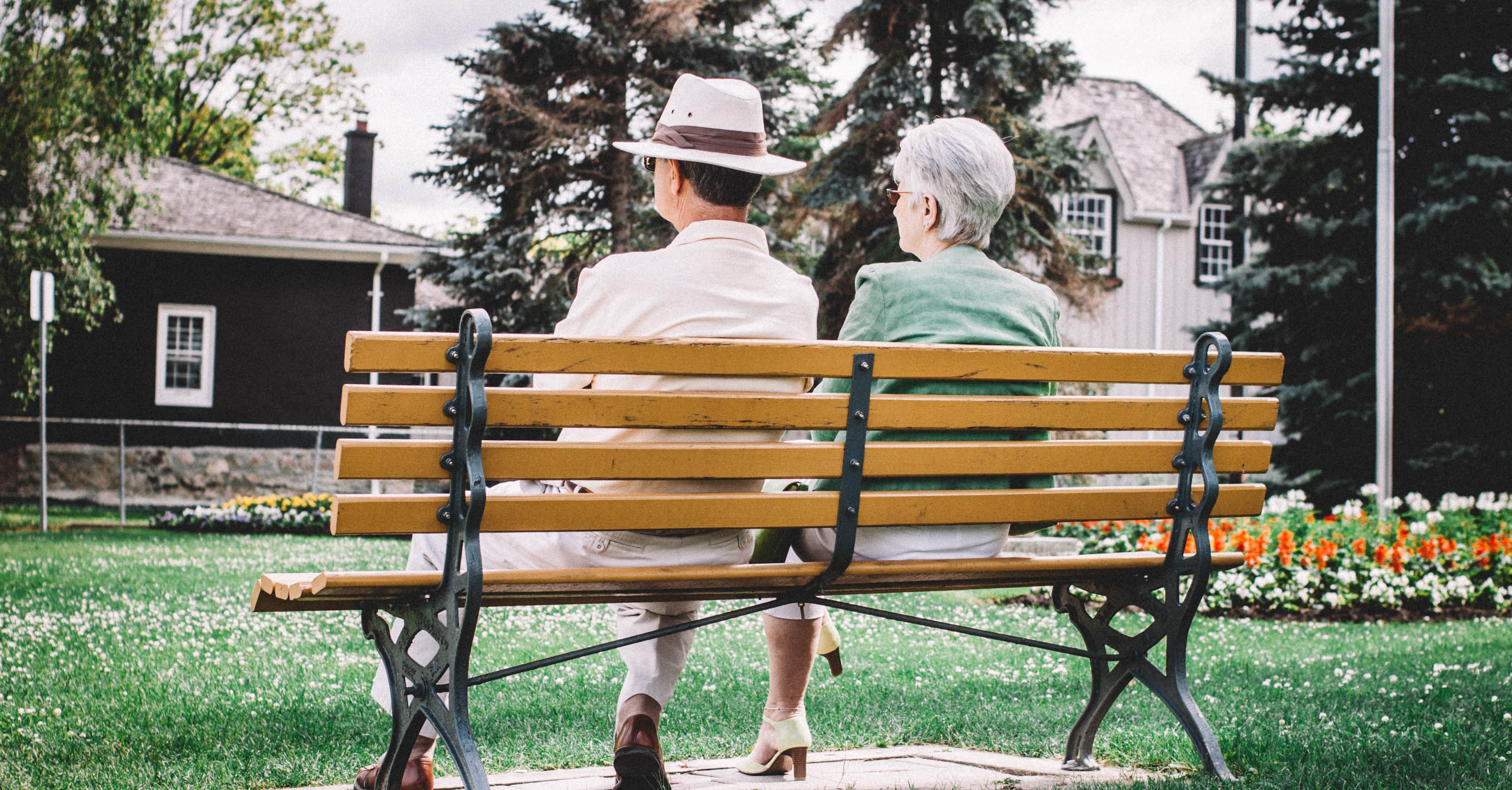 Two older adults sit on a bench in a park.