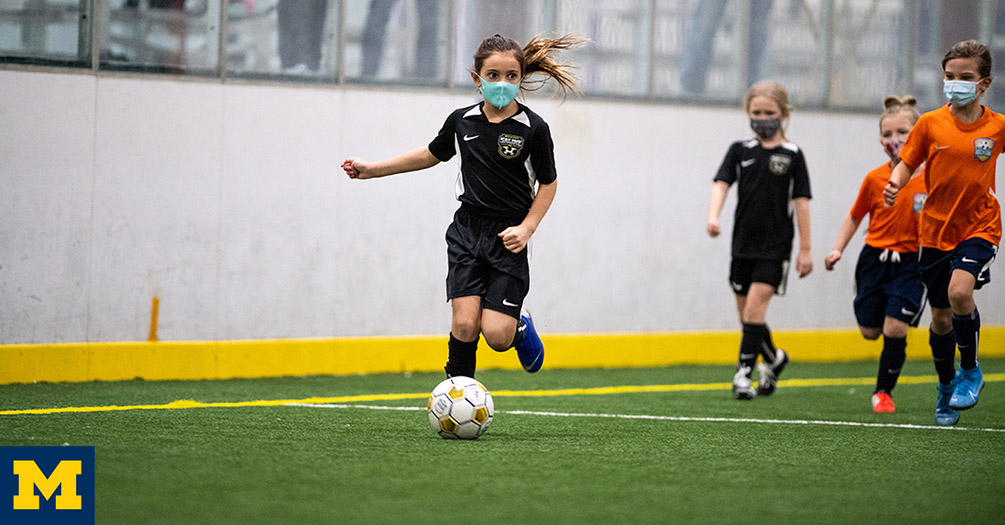 A group of children play soccer in an indoor arena with masks on.