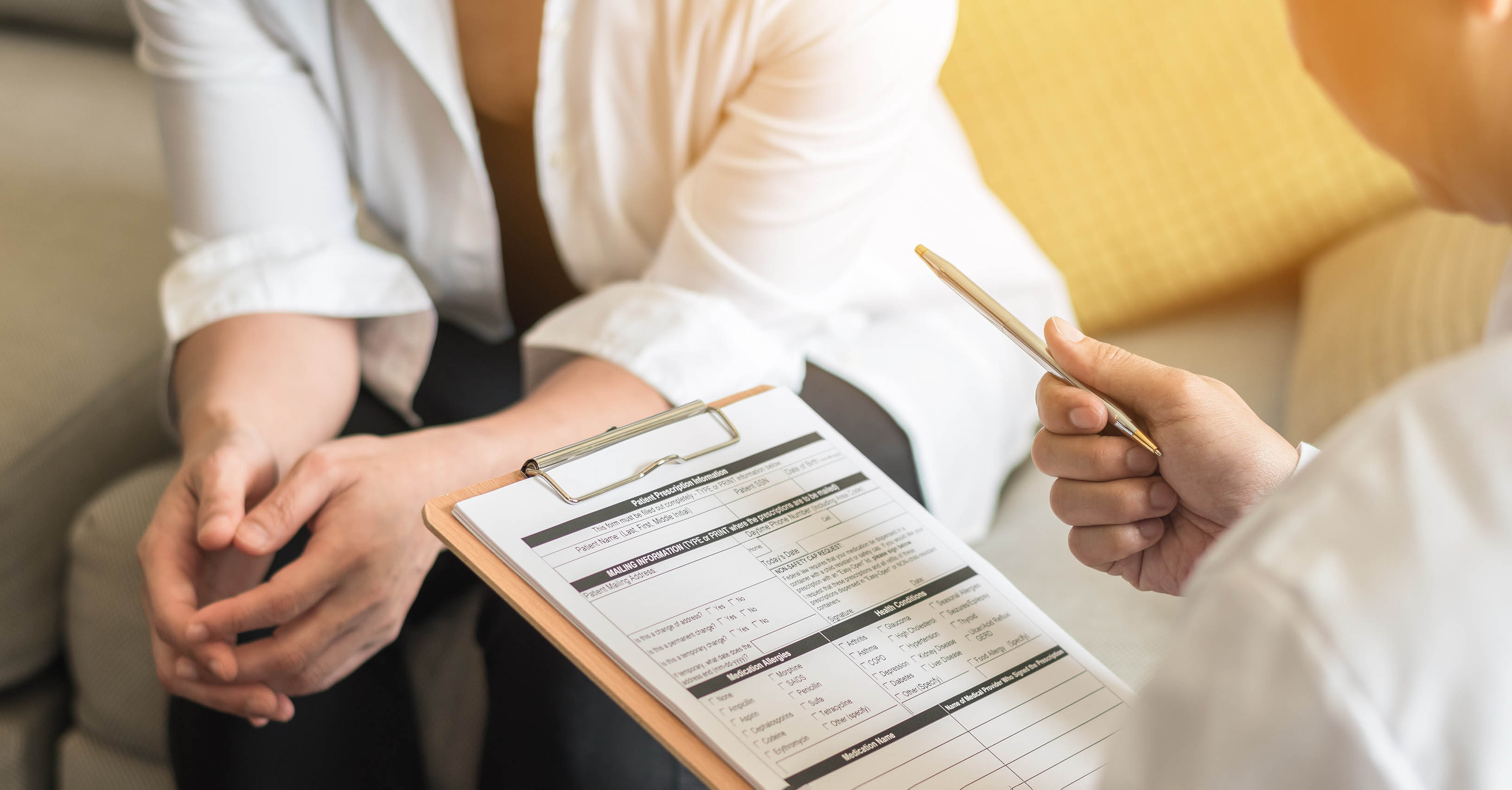 A female patient speaks with a medical professional who is holding a clipboard.