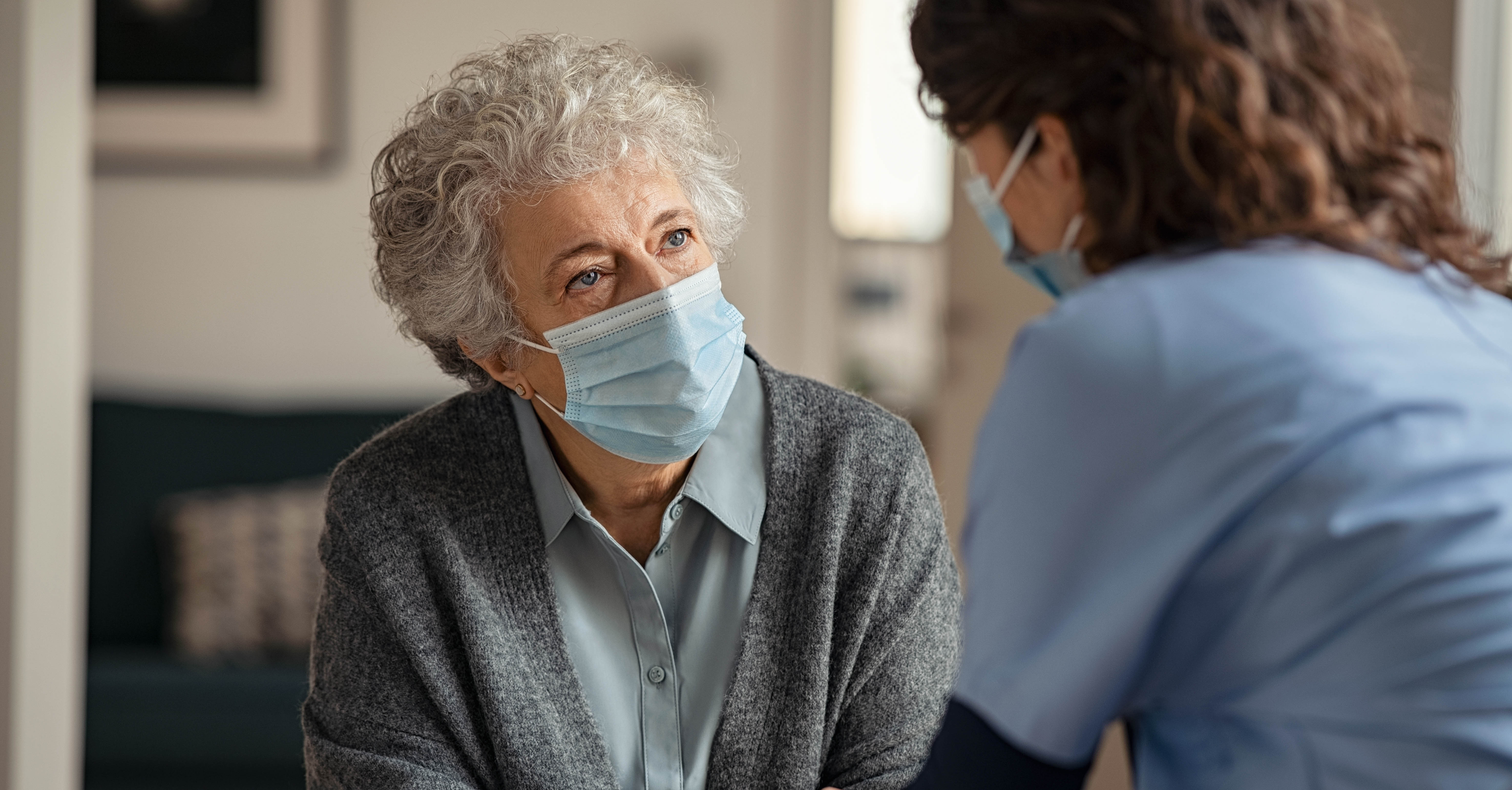 An elderly woman wearing a surgical mask speaks to a nurse.