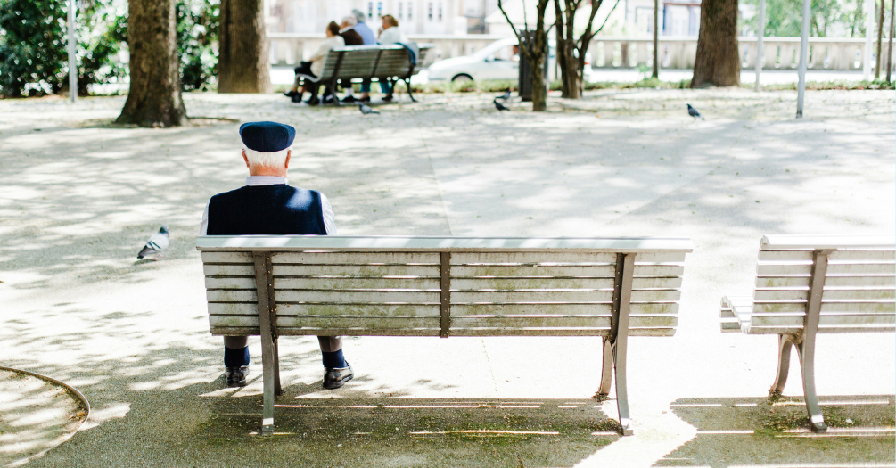 An older adult sits on a park bench alone.