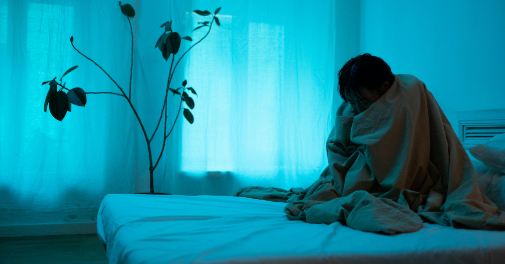 A person huddles in a blanket in their room, alone