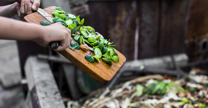 Hands sliding vegetable scraps from a cutting board into a compost bin