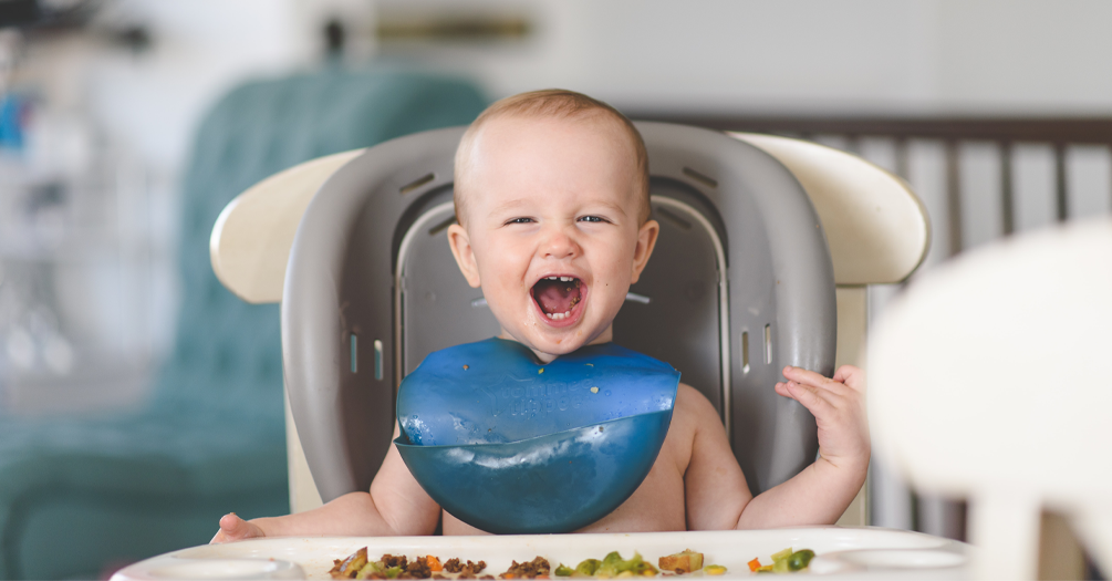 An infant in a high-chair opens his mouth wide as he eats food.