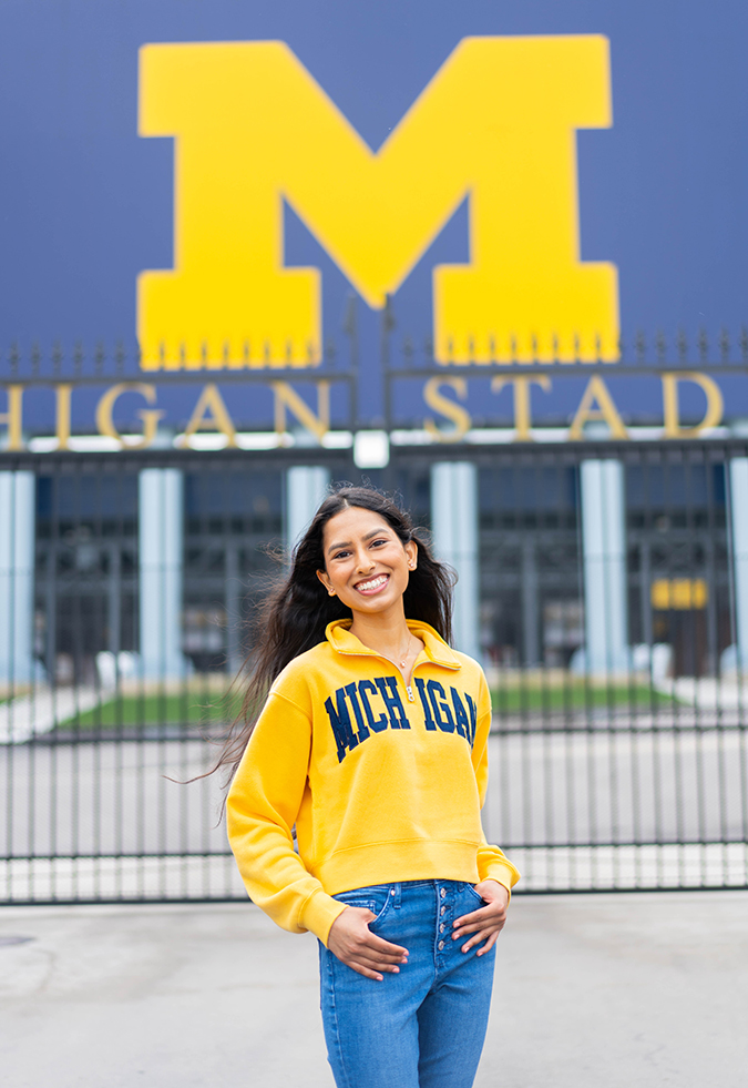 Aparna Reddy with a maize sheatshirt that reads "Michigan" in blue in front of the entrance to Michigan Stadium